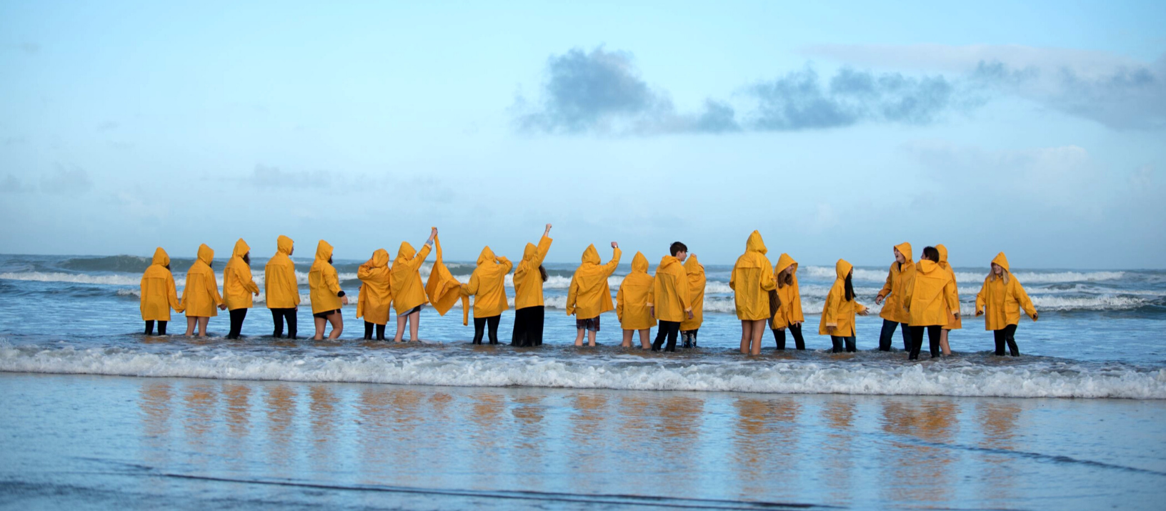 High Tide Don't Hide students standing in a line in yellow rain jackets holding hands in the air. All standing in the tide facing out to sea.